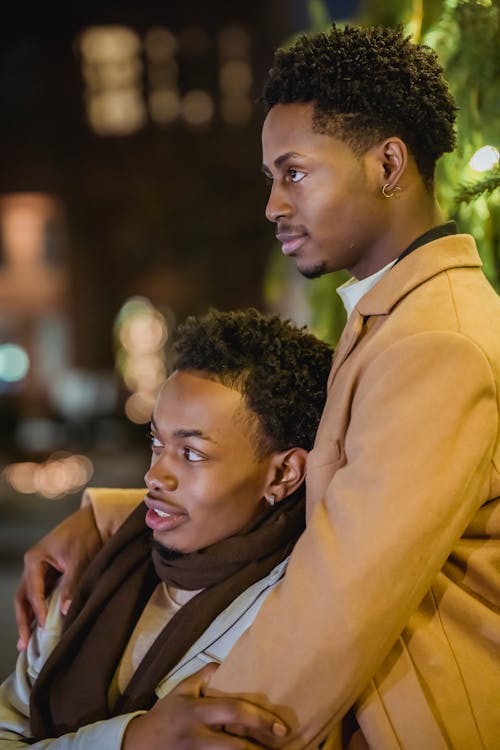 Side view of African American male couple in outerwear hugging gently and attentively looking away against lights