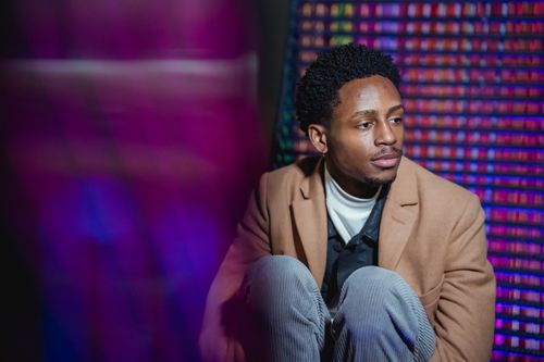 Free Peaceful African American male in coat embracing knees and looking away with serious glance Stock Photo