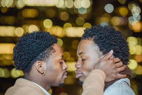 Free Side view of confident young African American homosexual men in warm clothes standing with closed eyes while embracing near glowing buildings in city street at night Stock Photo