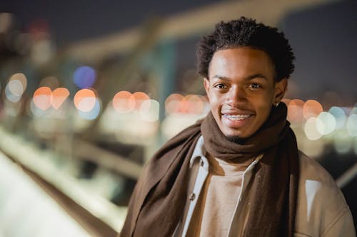 Positive African American male in trendy outfit looking away while standing on modern bridge against blurred modern in evening time