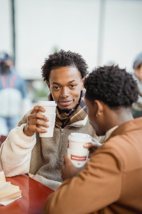 Content African American man with anonymous partner sitting at table with paper cups of hot beverages while spending time in cafe on blurred background