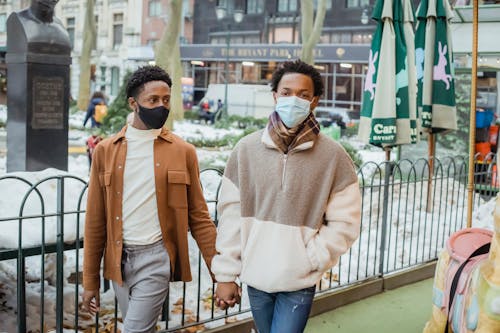 Gentle African American homosexual couple wearing casual wear in protective masks holding hands while strolling on street during coronavirus pandemic in city