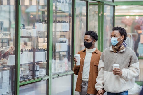 Free African American homosexual couple with takeaway beverages in medical masks holding hands while strolling on street near glass case of store in city Stock Photo