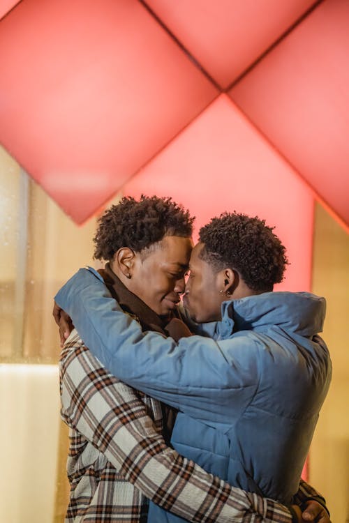 Side view of young ethnic gays with Afro haircuts in outerwear embracing in moment of kiss