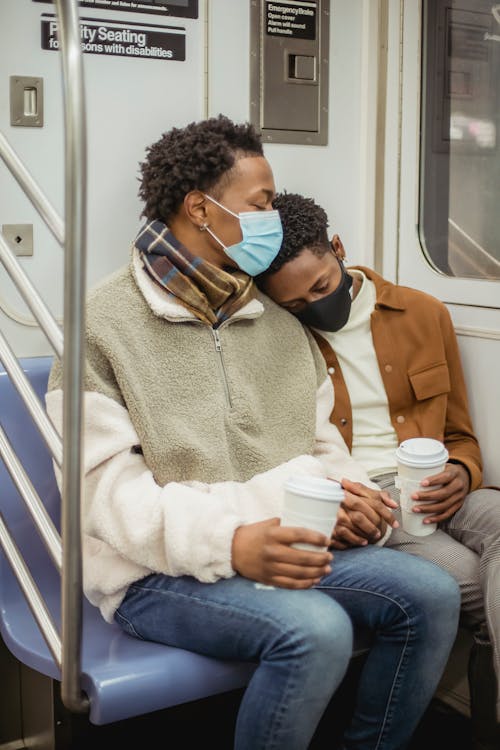 African American male passenger leaned on shoulder of homosexual boyfriend with hot beverage to go while commuting on subway
