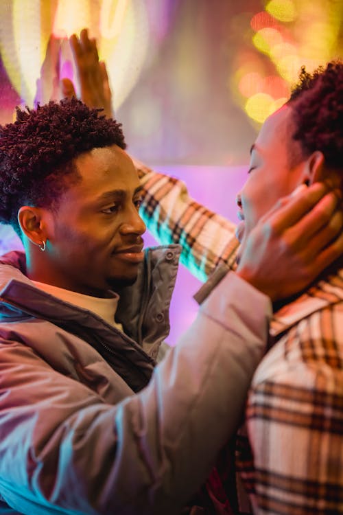 Side view of enamored young African American man in casual clothes smiling and touching face of boyfriend standing on illuminated city street