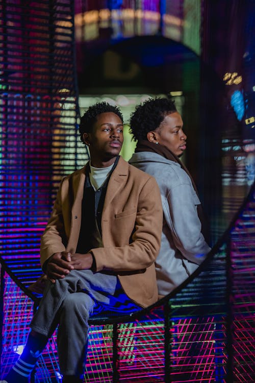 Trendy young black homosexual guys in stylish outfits sitting back to back on wicker hanging swing chair in illuminated room and looking away