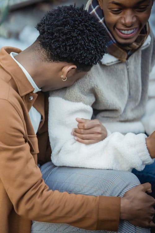 Unrecognizable cheerful young African American guy in trendy clothes holding arm of crop happy boyfriend and laughing while sitting together on city street