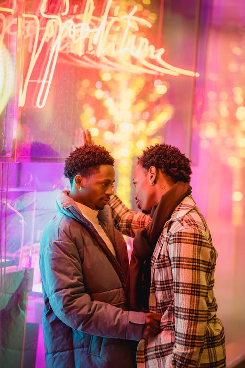 Side view of stylish young African American homosexual guys in warm clothes cuddling leaning on glass wall of modern building with neon illumination and looking at each other