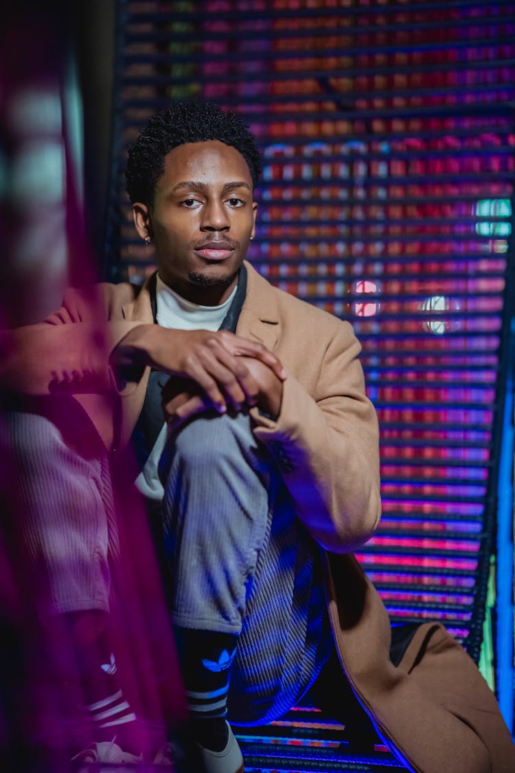 Stylish Young Black Guy Relaxing On Wicker Chair In Room With Neon Lights