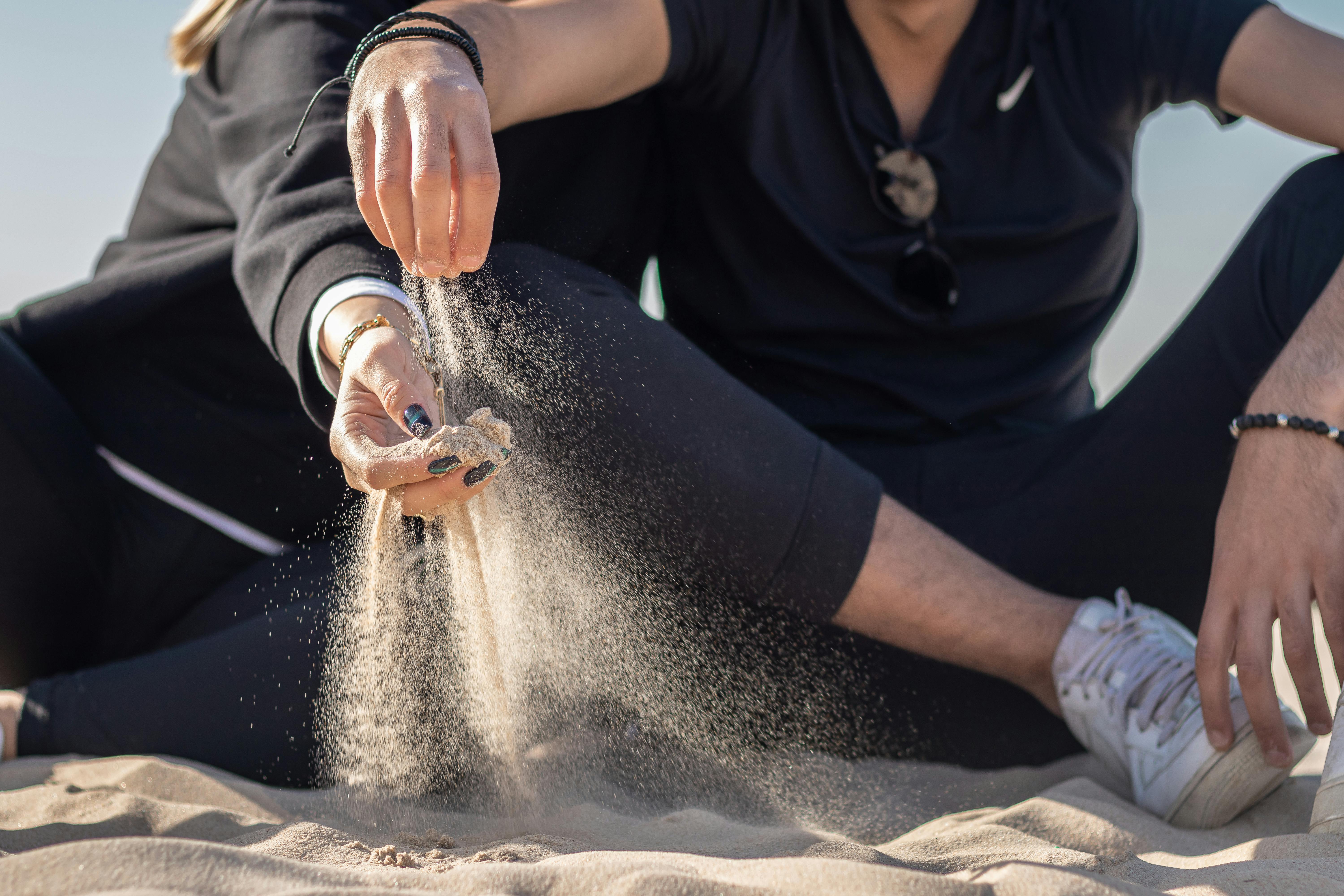 Free From below of crop anonymous couple sitting on sandy beach legs crossed together and flowing sand through fingers under blue sky wealth, problem-solving, financial success, value creation