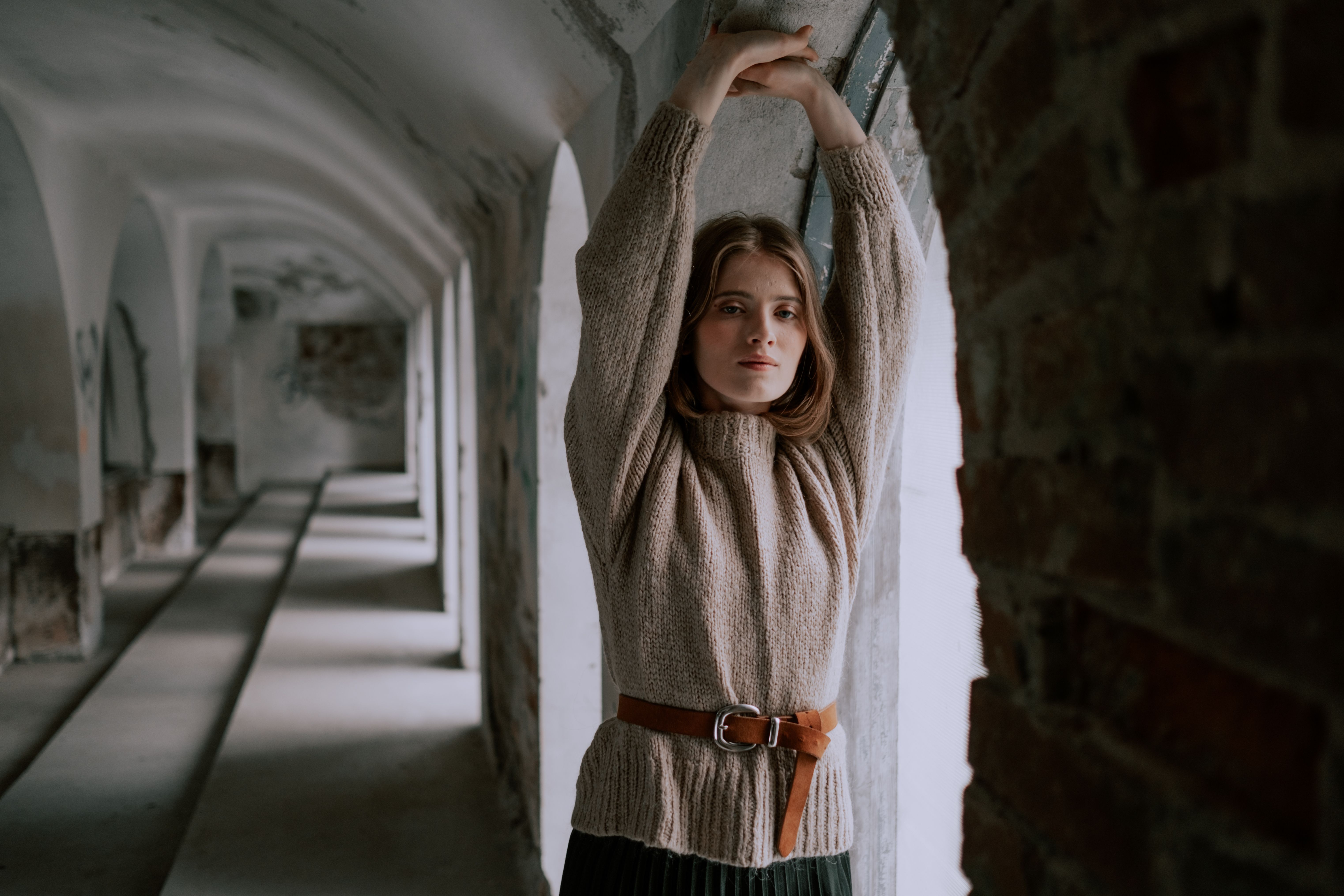 Woman in cozy sweater stretches by arched window in historic interior setting.