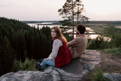 Free A couple sits on a rock overlooking a serene forest and river landscape at sunset. Stock Photo