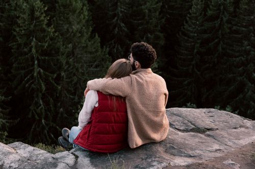 Free A couple sits embracing on a rocky ledge facing a dense forest. Capture the essence of nature and togetherness. Stock Photo