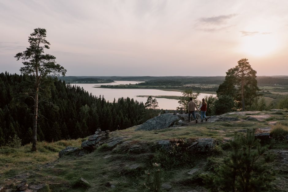 A couple enjoys a serene walk on a rocky cliff overlooking a tranquil lake and forest during sunset.