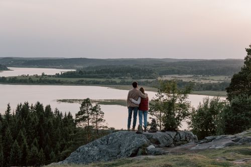 Free A couple standing on rocks, embracing, overlooking a serene lake and forest at sunset. Stock Photo