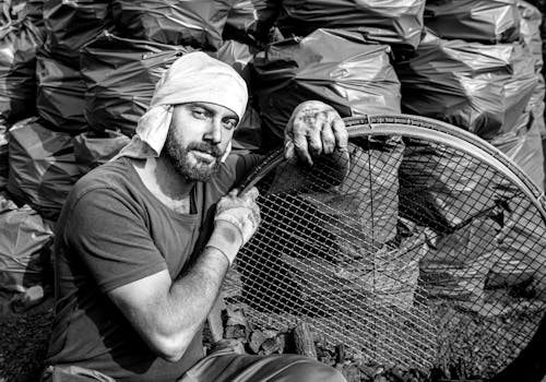 Black and White Photo of Man Sitting among Trash Bags Holding Circular Crate