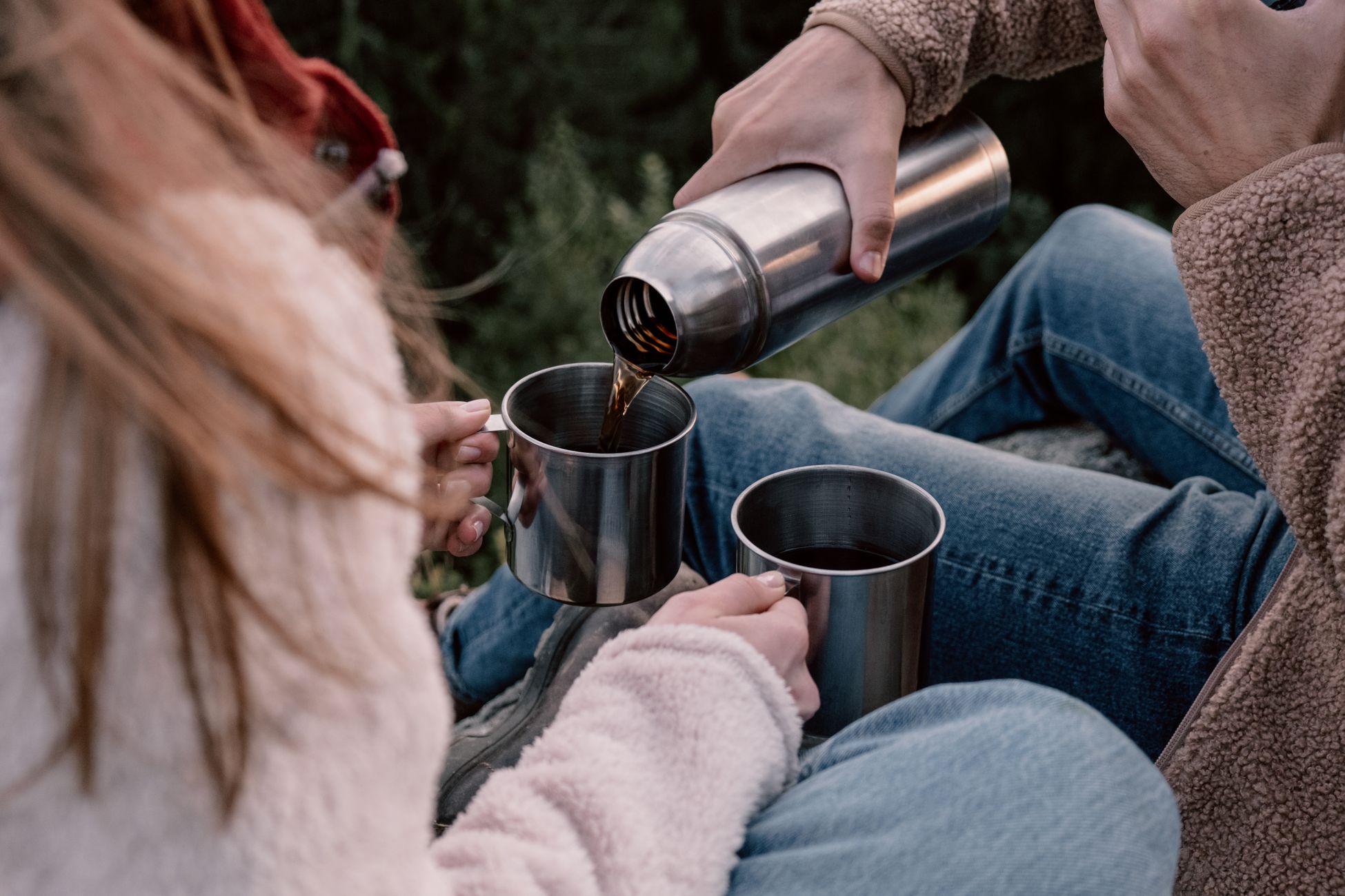 A couple pours hot coffee from a thermos into mugs while seated outdoors.