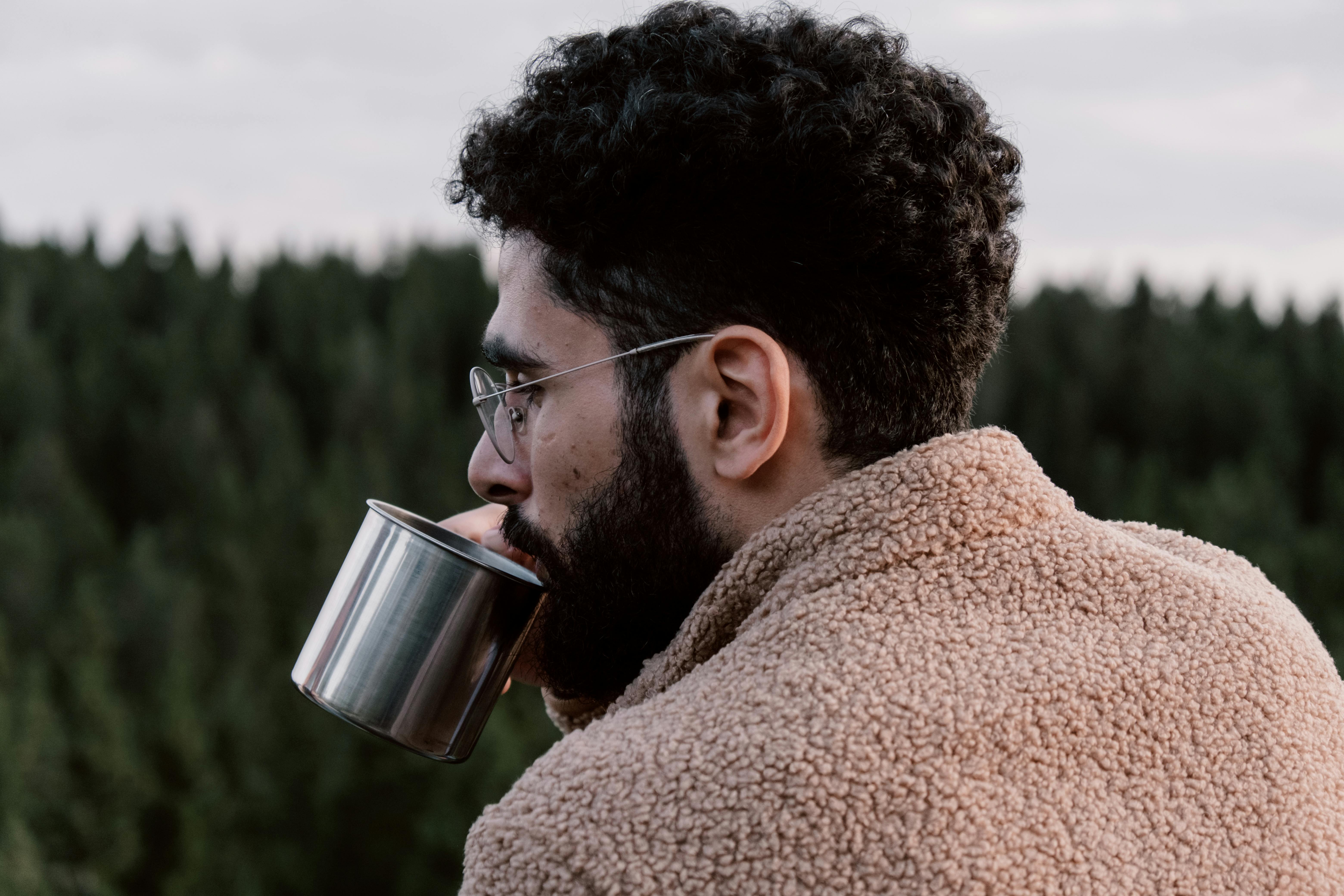 A man with curly hair and glasses enjoys a hot drink outdoors, wrapped in a cozy sweater.