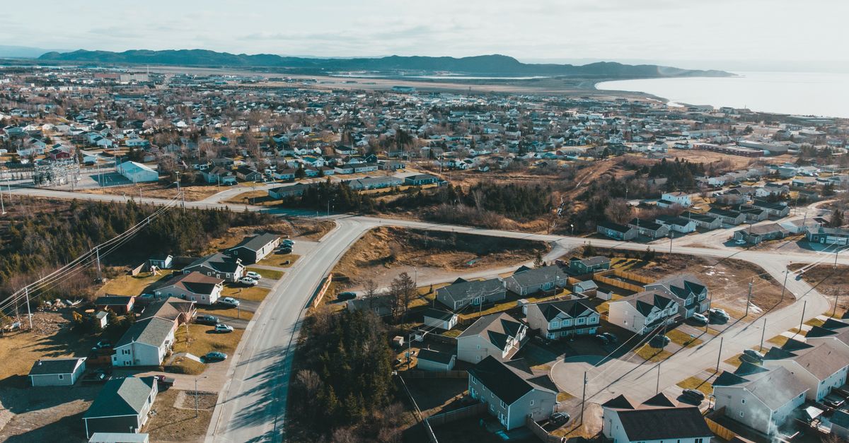 Drone view of coastal settlement with small houses and asphalt road near sea and mountain on background under light blue sky on sunny day in autumn