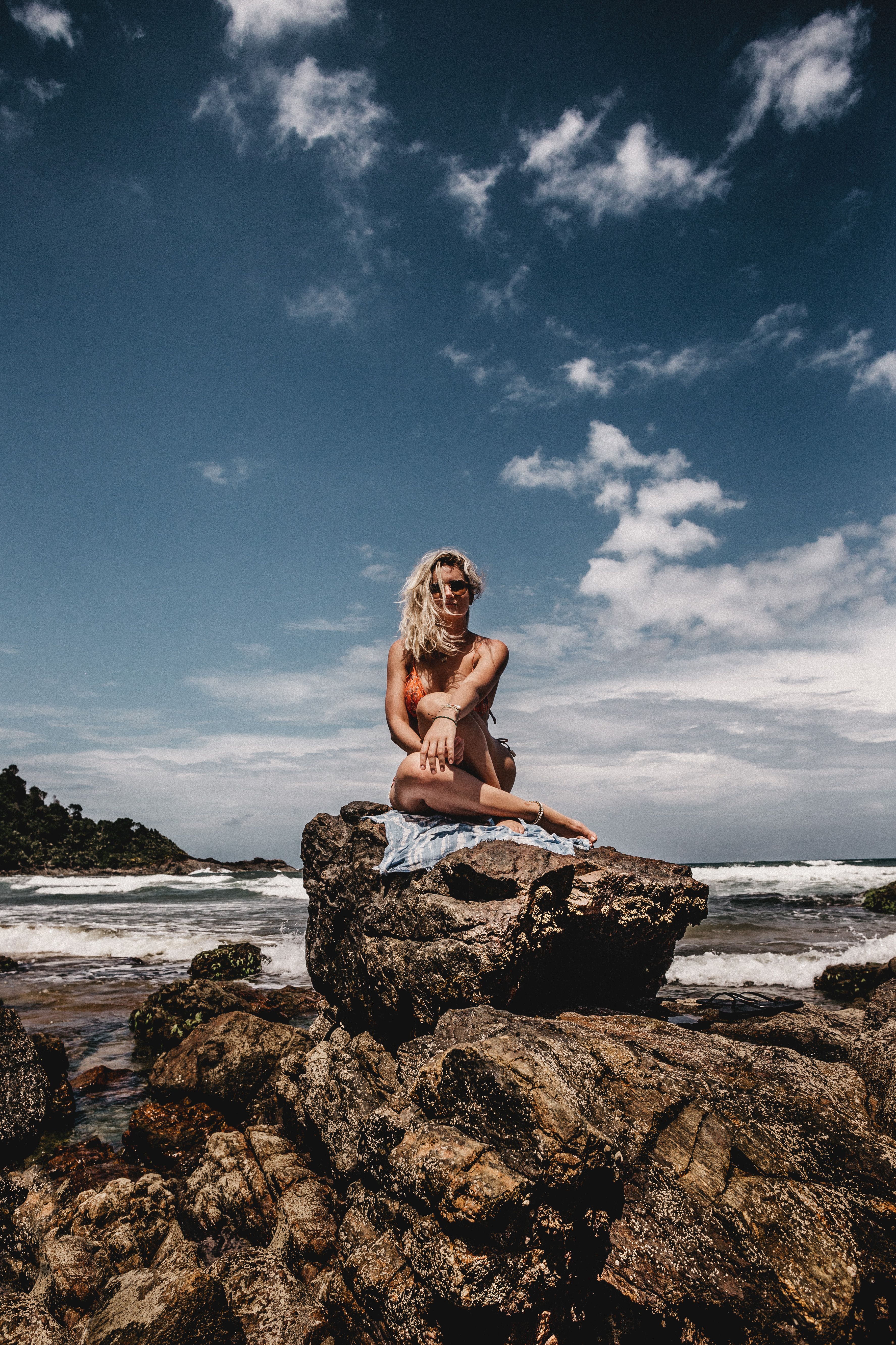 Tourist resting on rough rocky formation on coast against stormy ocean under blue cloudy sky during summer vacation