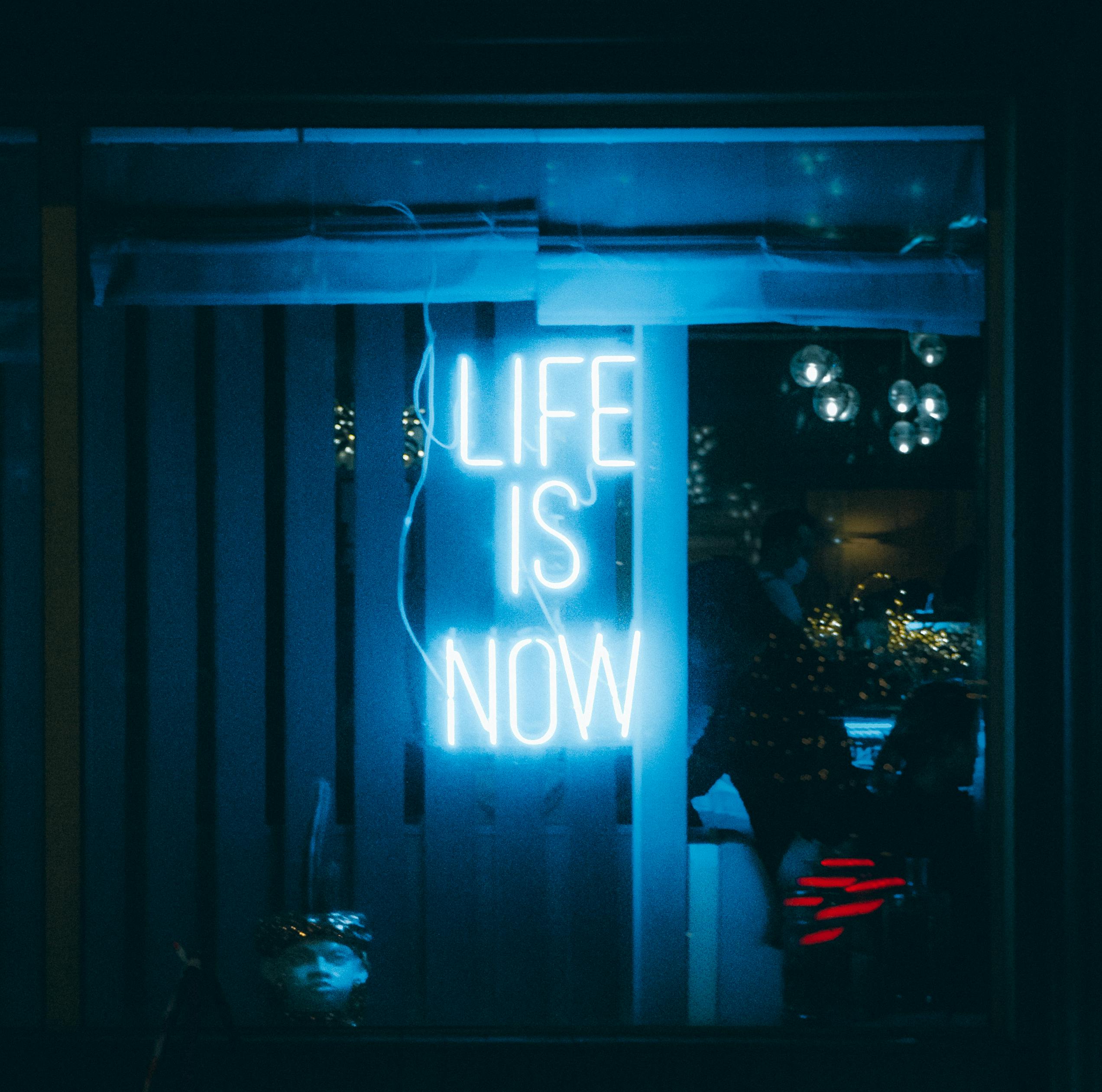 A glowing neon sign displaying 'Life is Now' seen from outside a bar at night.