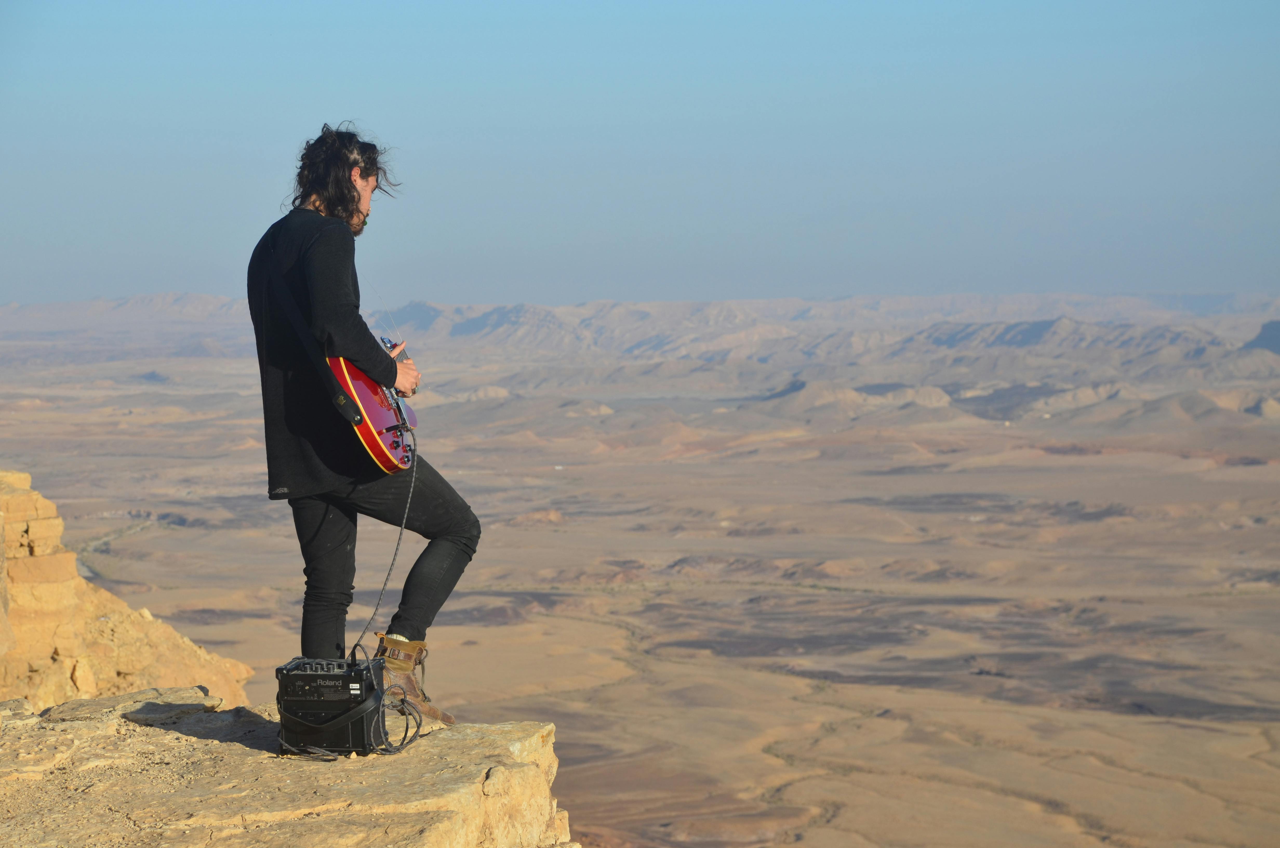 Musician playing electric guitar on a cliff with a vast desert landscape.
