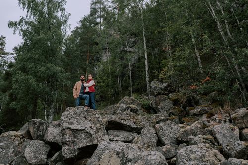 Free A couple stands embracing on a rocky terrain surrounded by lush forest, capturing a serene outdoor moment. Stock Photo