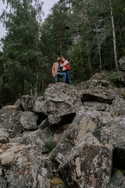Free A couple stands on large rocks surrounded by a dense forest, conveying a sense of adventure and tranquility. Stock Photo