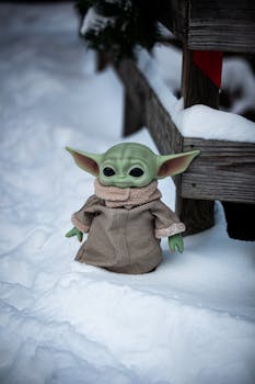 Grogu toy dressed warmly, posing on snow-covered ground for a winter-themed photo.