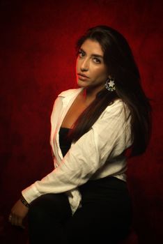Pensive young ethnic female wearing white shirt and black clothes with earrings while sitting in bright studio on red background and looking at camera