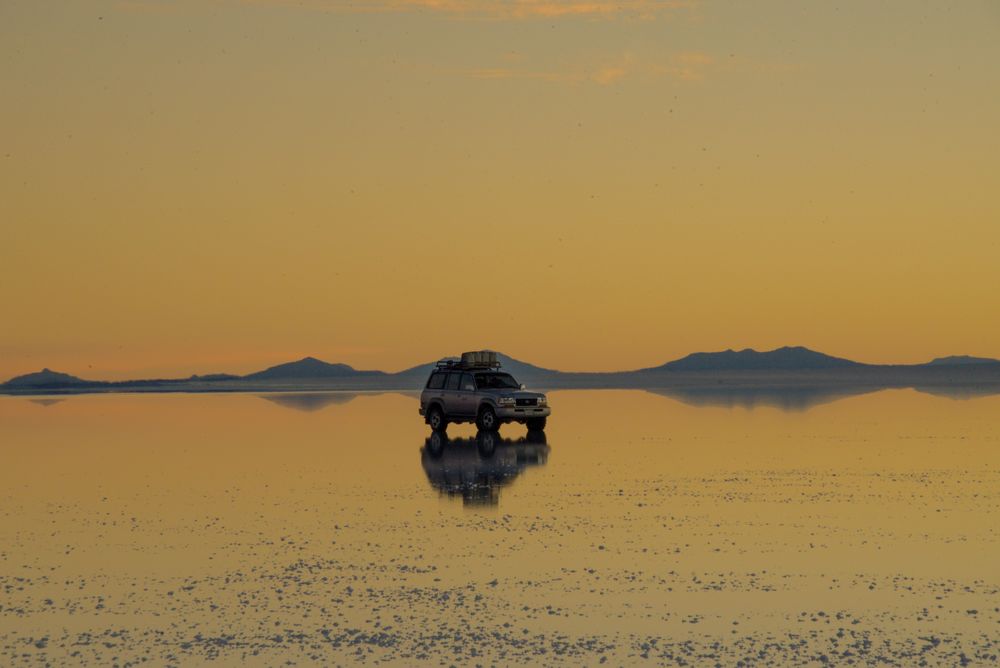 Vehículo 4x4 sobre el espejo del Salar de Uyuni al ocaso con volcanes al fondo