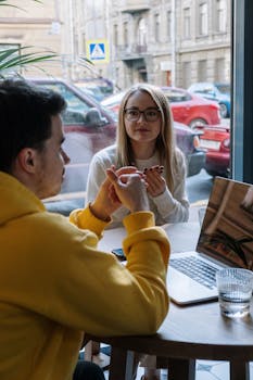 A man and woman having a casual discussion in a modern cafe setting with laptops.