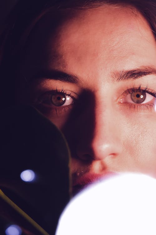 Closeup of crop young tranquil lady looking at camera thoughtful through glass window reflecting glowing lights in dark room