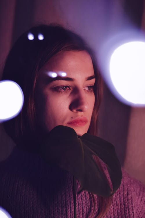 Through glass of young ethnic melancholic female with brown hair in sweater looking away sadly while standing near glowing lamps in dark room with fresh Monstera deliciosa plant in hand