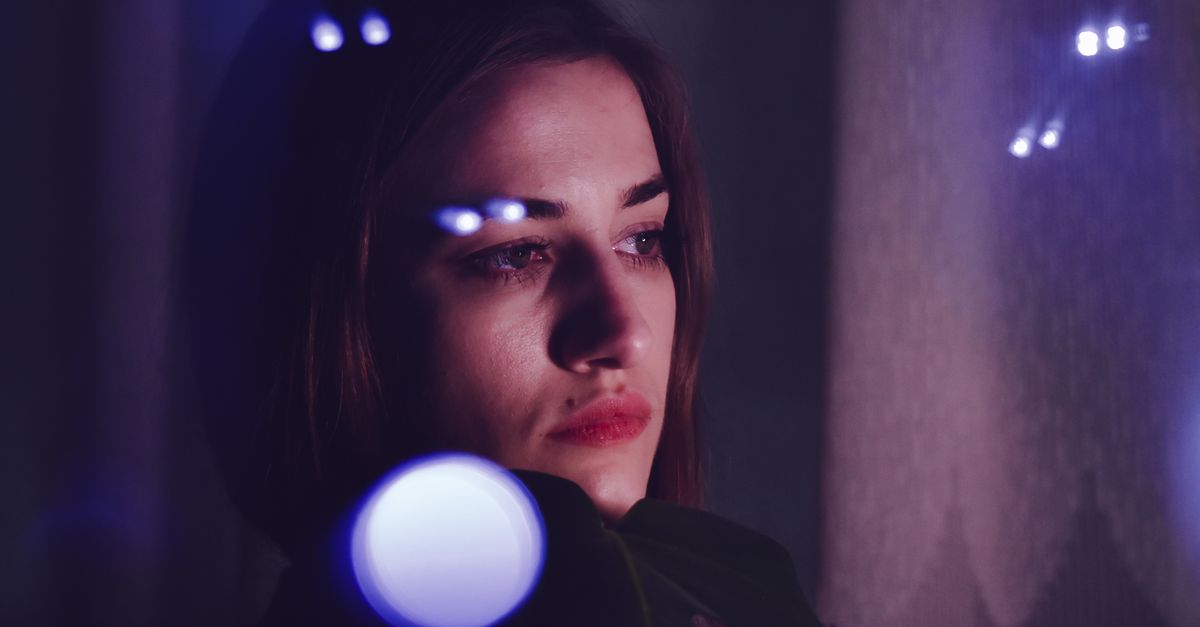 Through glass of young upset ethnic female with long brown hair looking away thoughtfully while resting in dark room near window reflecting light glares