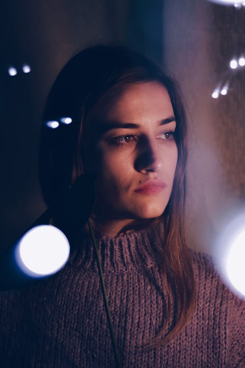 Through glass with light glares of serious young ethnic female millennial with long hair in warm sweater standing near window in dark room and looking away