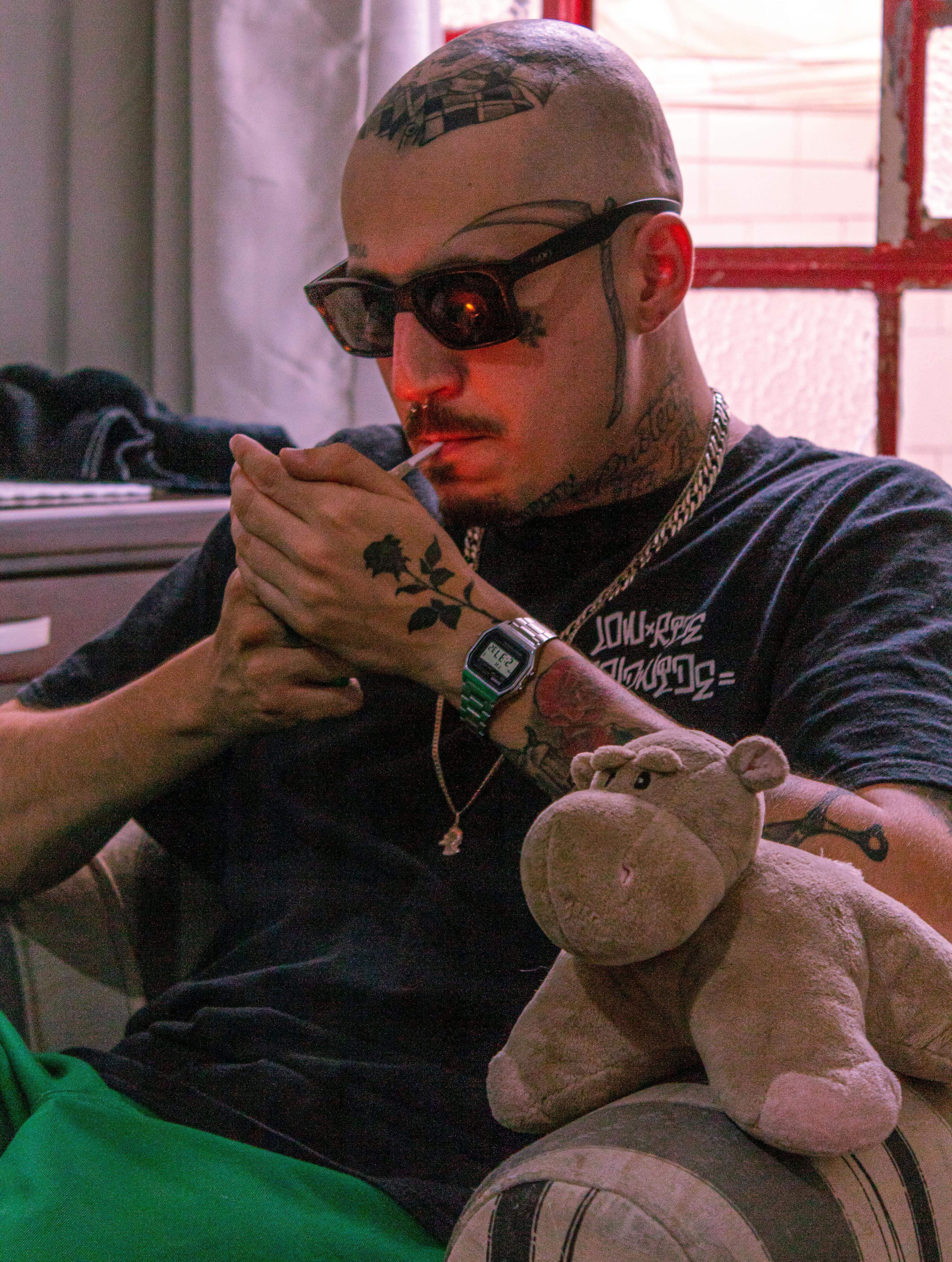 Serious male with tattoos and sunglasses wearing chain smoking cigarette while sitting in armchair with toy in living room at home
