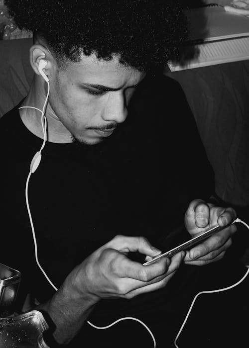 Black and white of concentrated ethnic male with earphones playing game on modern cellphone while sitting on sofa at home