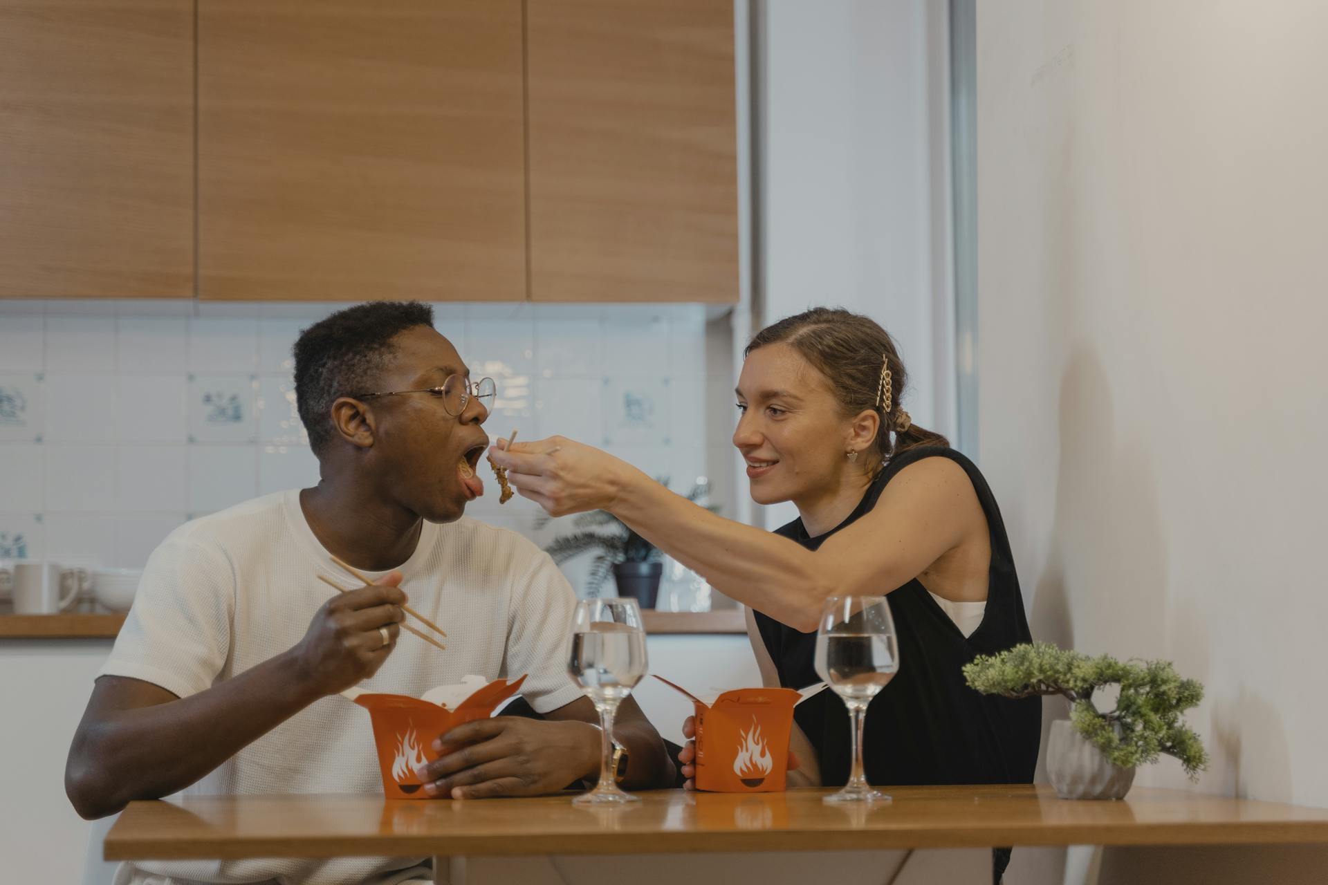 An interracial couple sharing a meal with chopsticks at a cozy dinner table. Food delivery scene.