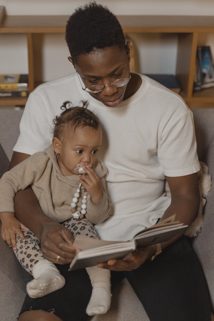 A Man Sitting On Couch Reading Book To A Baby With Pacifier
