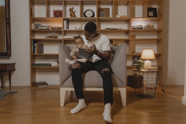 A Man Sitting On Gray Chair With A Baby Holding A  Book