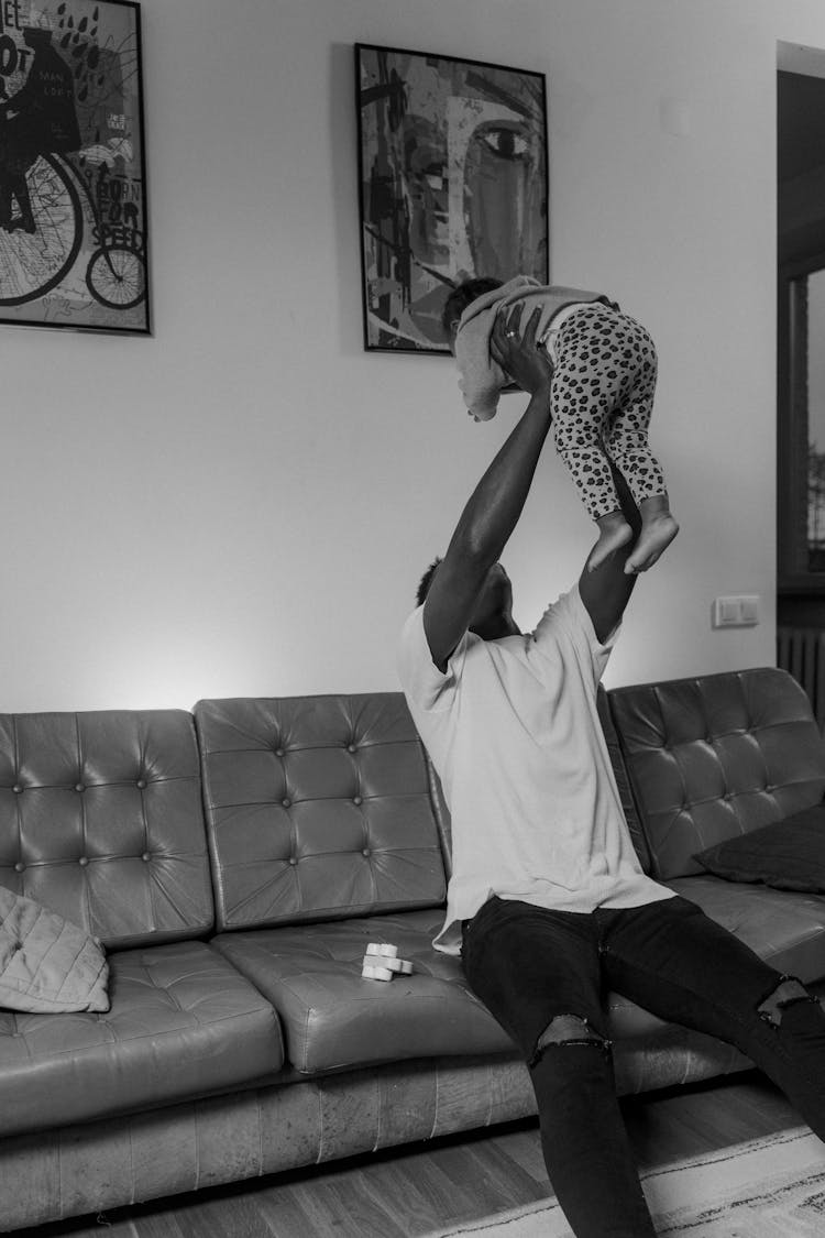 Grayscale Photo Of A Man Sitting On Couch Lifting A Baby