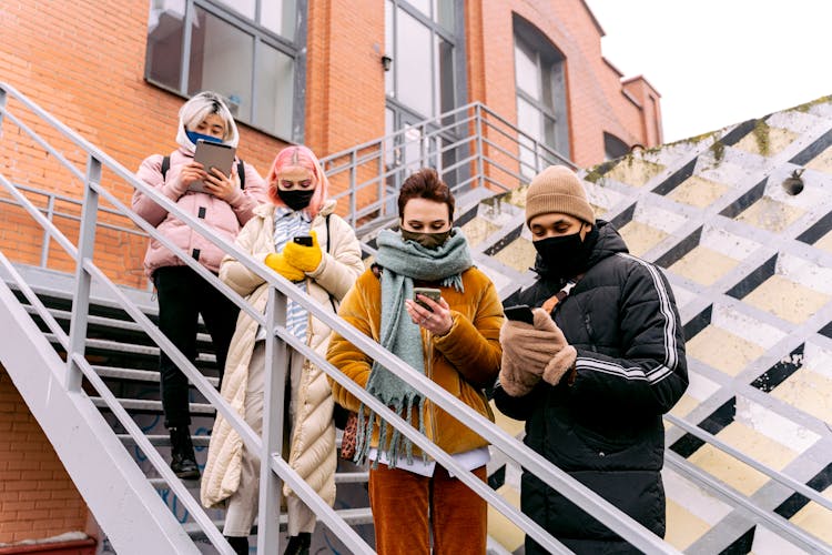 Group Of Young People Standing On Stairs, Using Phones And Wearing Face Masks 