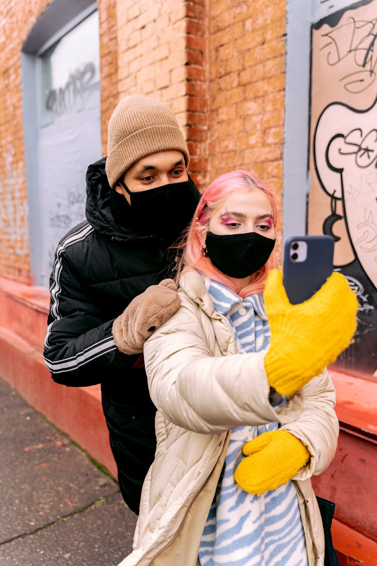 Man And Woman In Face Mask And Winter Clothing Taking A Selfie On A Sidewalk 