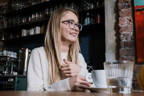Free A cheerful young woman with glasses smiling while holding a cup in a cozy cafe setting. Stock Photo