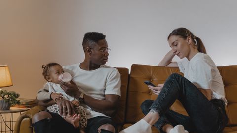Parents relaxing on a sofa while feeding their baby, enjoying family time with a cellphone.