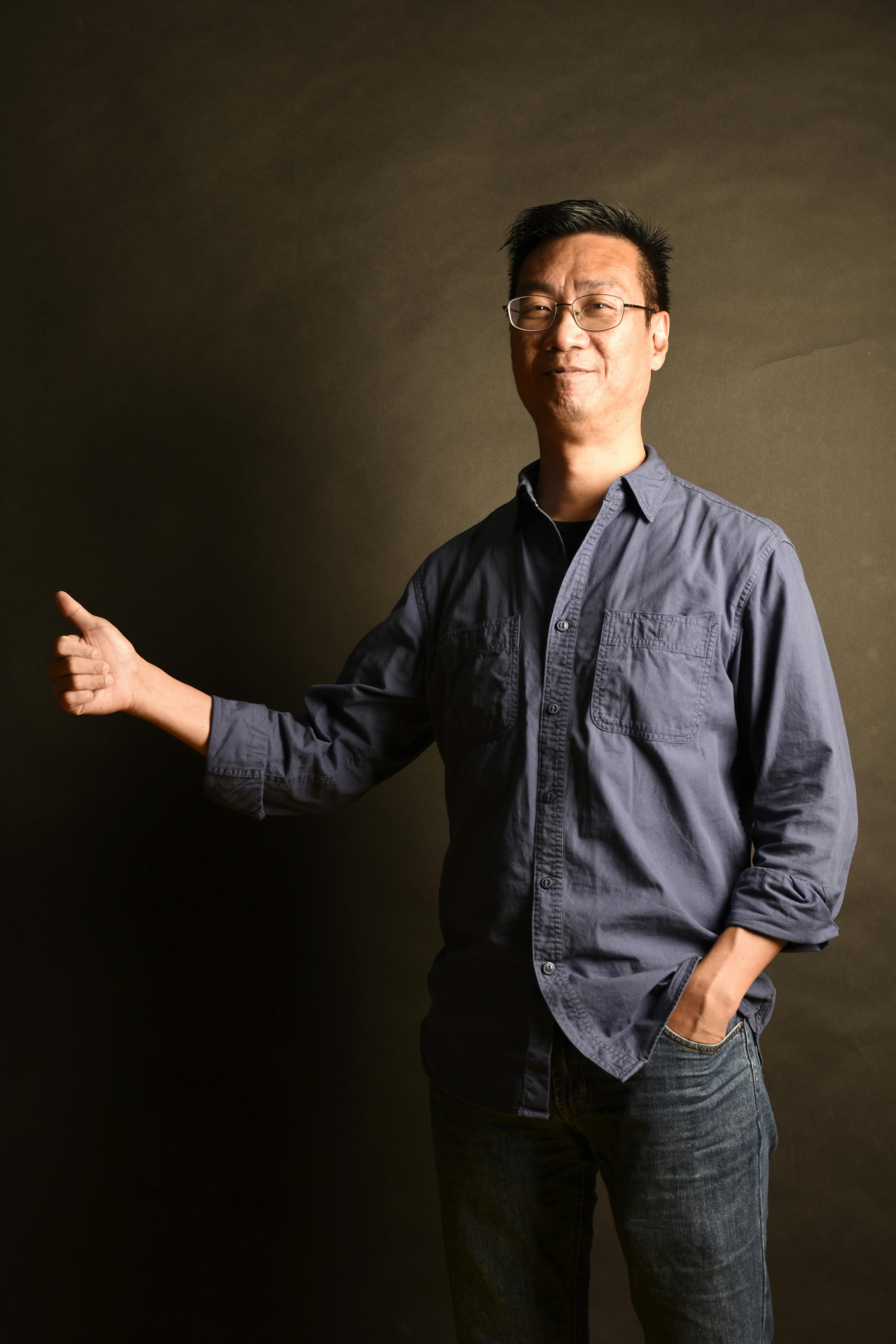 Asian man in jeans and shirt posing with thumbs up gesture in a studio setting.