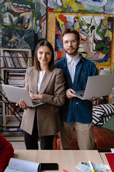 Two creative professionals holding laptops in a colorful art studio setting.