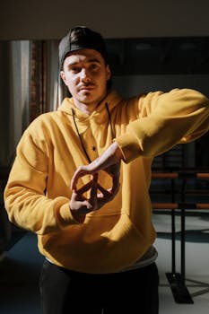 Portrait of a man in a yellow hoodie performing a peace sign gesture indoors.